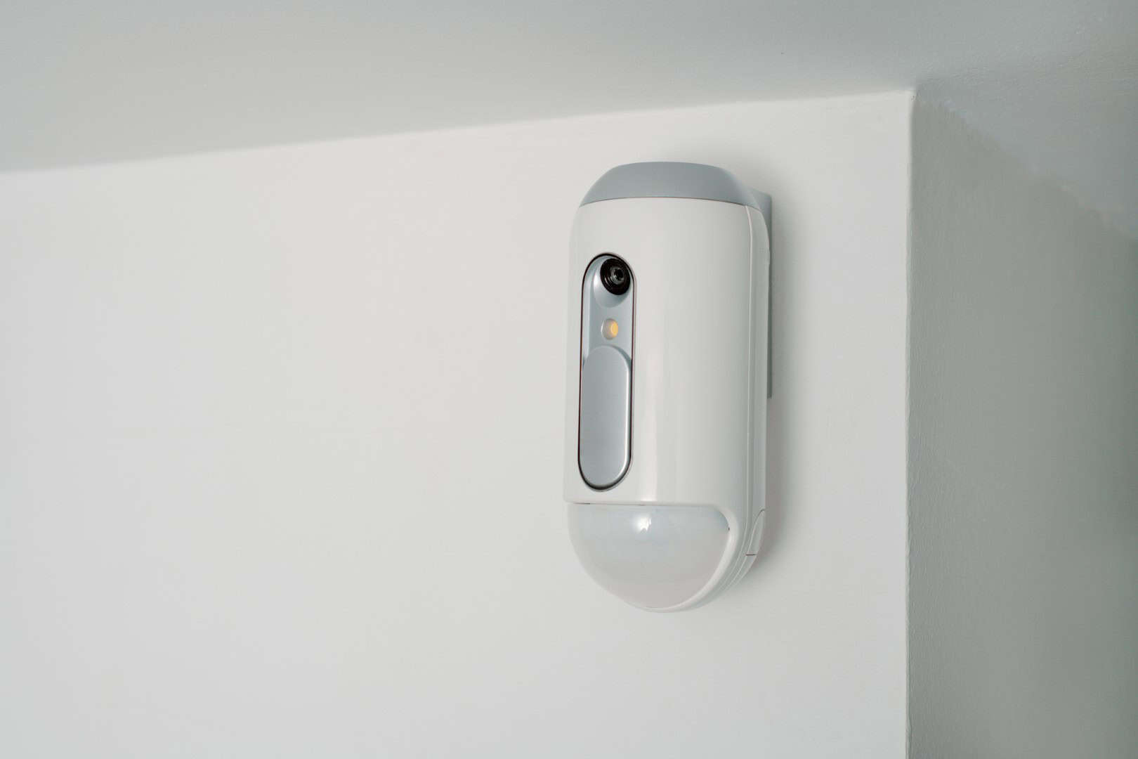 a motion detector equipped with a camera on a white wall, indoors, as part of a burglar alarm system